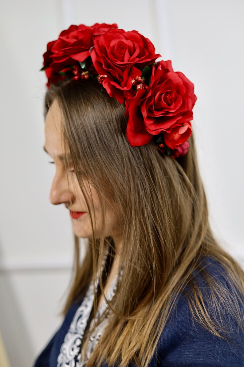 K&ouml;nnte beinhalten: Ein Blumenkranz mit gro&szlig;en, tiefroten Rosen. Der Kopfschmuck besteht aus realistisch aussehenden Rosen und kleinen roten Beeren. Die Person tr&auml;gt ein dunkelblaues Oberteil. Die roten Rosen sind ein Blickfang.