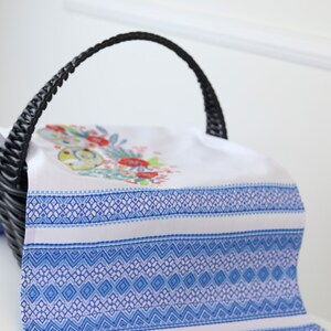 May include: White and blue patterned decorative towel with floral embroidery, draped over a black wicker basket. The towel features a detailed blue geometric design and a floral motif with red and green accents.