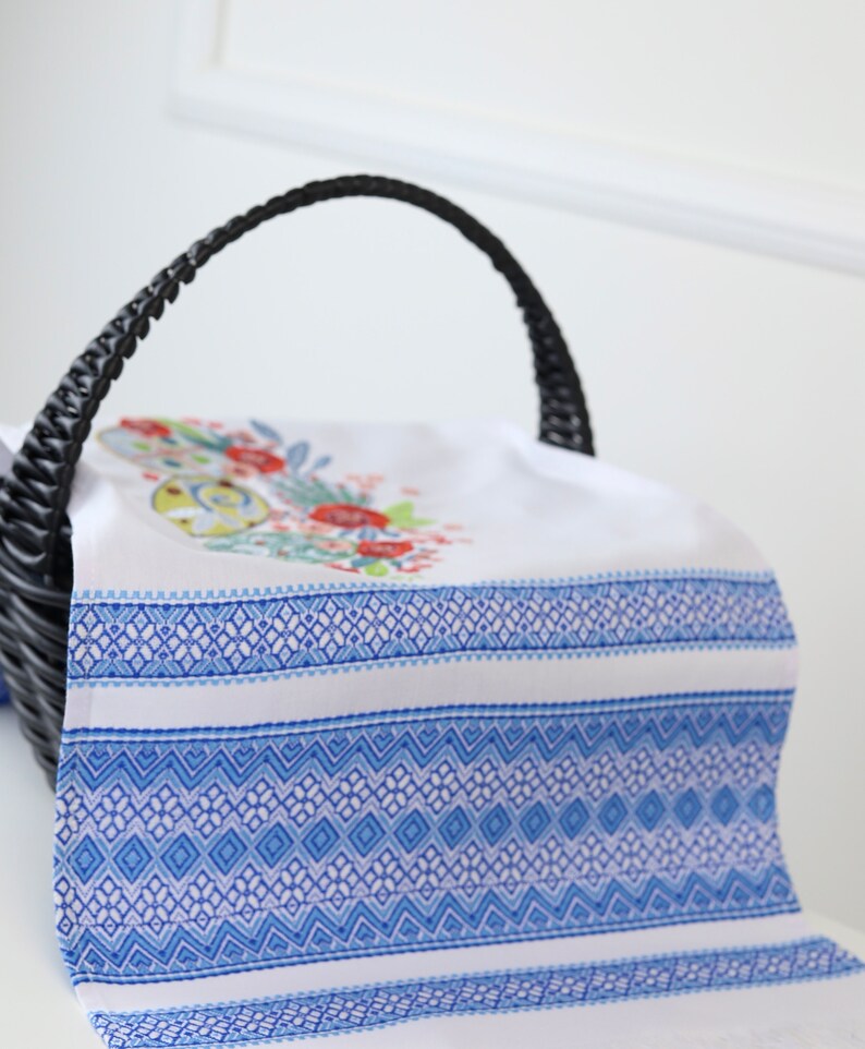 May include: White and blue patterned decorative towel with floral embroidery, draped over a black wicker basket. The towel features a detailed blue geometric design and a floral motif with red and green accents.
