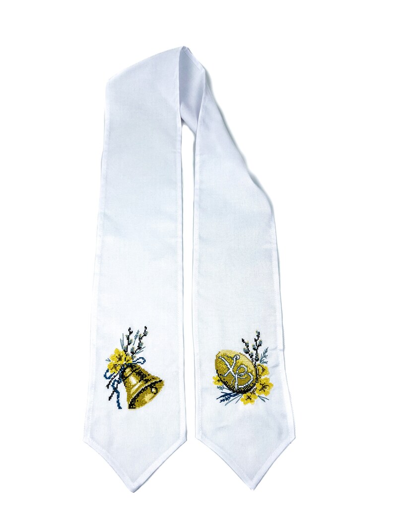 K&ouml;nnte beinhalten: Wei&szlig;er Stoffschal mit gestickten Ostermotiven. Eine Seite zeigt eine goldene Glocke mit gelben Blumen und gr&uuml;nen Zweigen. Die andere Seite hat ein gelbes Ei mit den kyrillischen Buchstaben 'XB' und gelben Blumen.