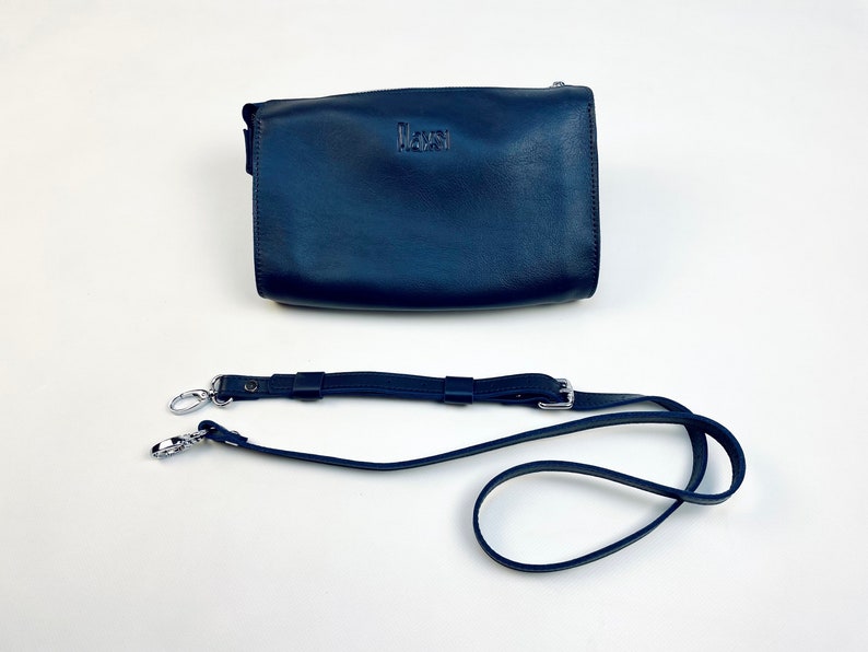May include: A dark blue leather crossbody bag with a silver strap and a silver clasp. The bag has the word "MAURI" embossed on the front.