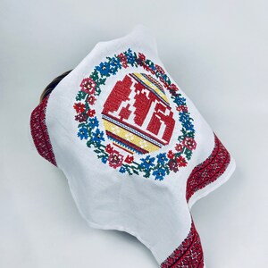 Pu&ograve; includere: Asciugamano da cucina in lino bianco con una ghirlanda di fiori ricamata in rosso e blu e un uovo di Pasqua rosso, giallo e blu al centro.