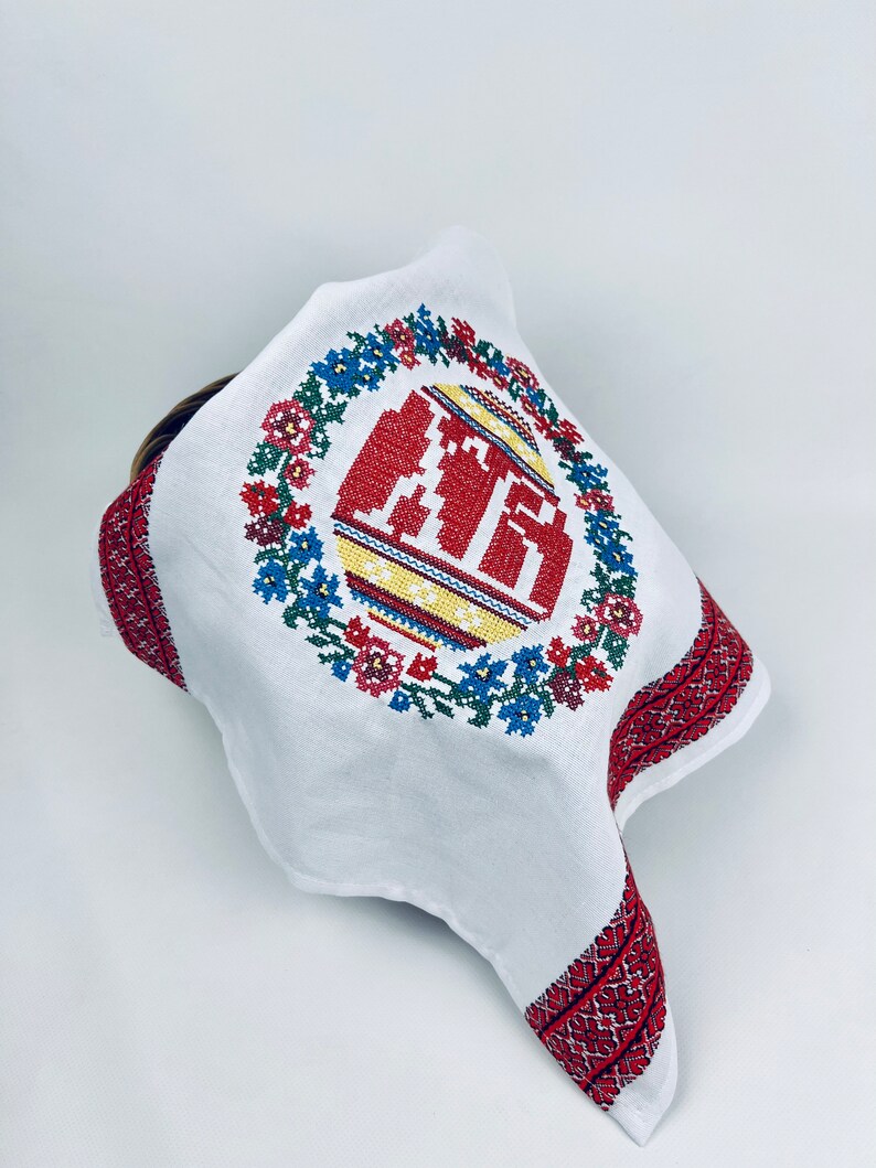 Pu&ograve; includere: Asciugamano da cucina in lino bianco con una ghirlanda di fiori ricamata in rosso e blu e un uovo di Pasqua rosso, giallo e blu al centro.