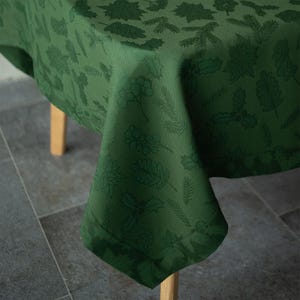 Op de afbeelding: Een groene tafelkleed met een herhalend patroon van bladeren en bessen. Het tafelkleed is over een houten tafel gedrapeerd, waarbij de hoek van de stof zichtbaar is. De stof heeft een subtiele glans.