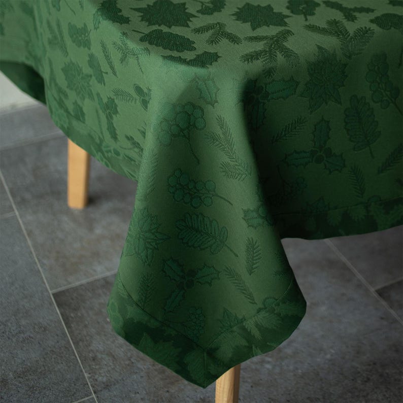 Op de afbeelding: Een groene tafelkleed met een herhalend patroon van bladeren en bessen. Het tafelkleed is over een houten tafel gedrapeerd, waarbij de hoek van de stof zichtbaar is. De stof heeft een subtiele glans.