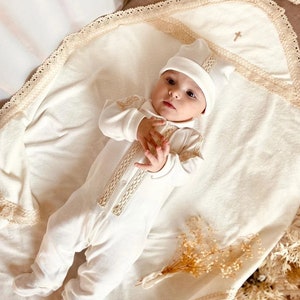 May include: A white baby onesie with a lace trim and a cross embroidered on the front. The baby is wearing a white hat and is lying on a white blanket with lace trim.