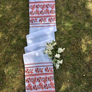 May include: A white embroidered table runner featuring a floral pattern in red and black. The runner is folded on green grass, with white flowers. The embroidered section is approximately 30 cm wide.