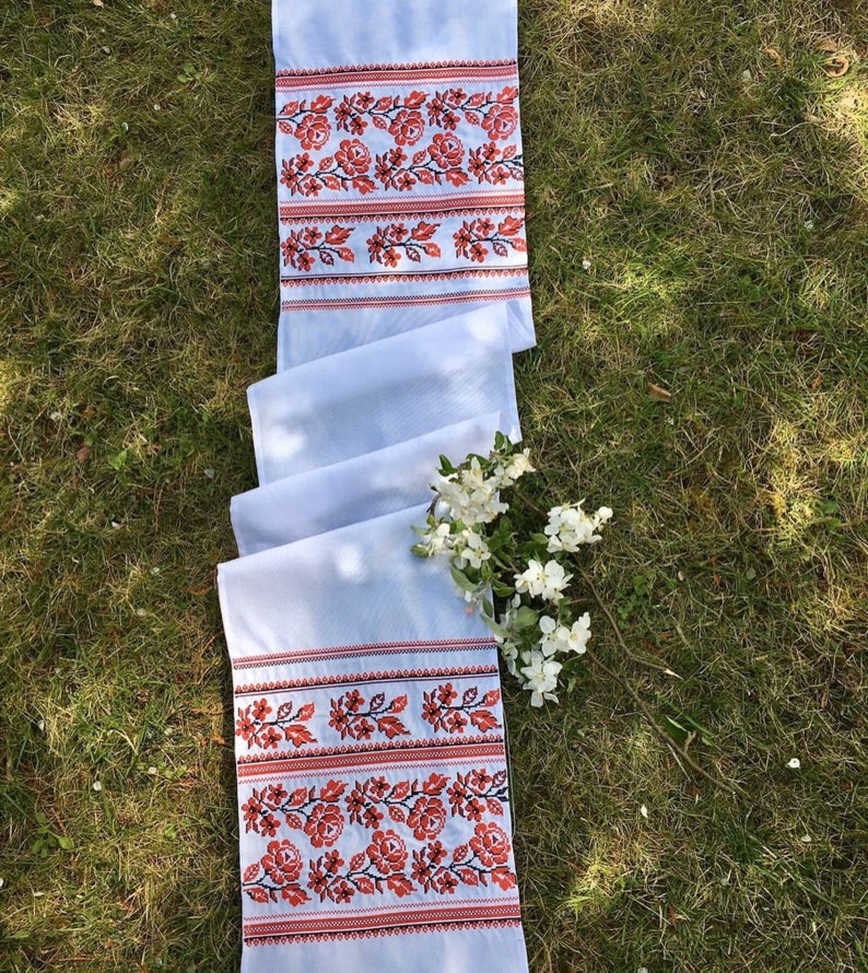 May include: A white embroidered table runner featuring a floral pattern in red and black. The runner is folded on green grass, with white flowers. The embroidered section is approximately 30 cm wide.