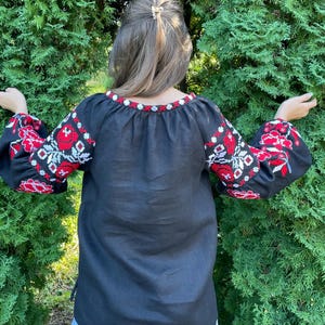 Puede incluir: Una camisa de lino negra con flores bordadas rojas y blancas en las mangas. La camisa tiene un cuello redondo y un corte holgado.