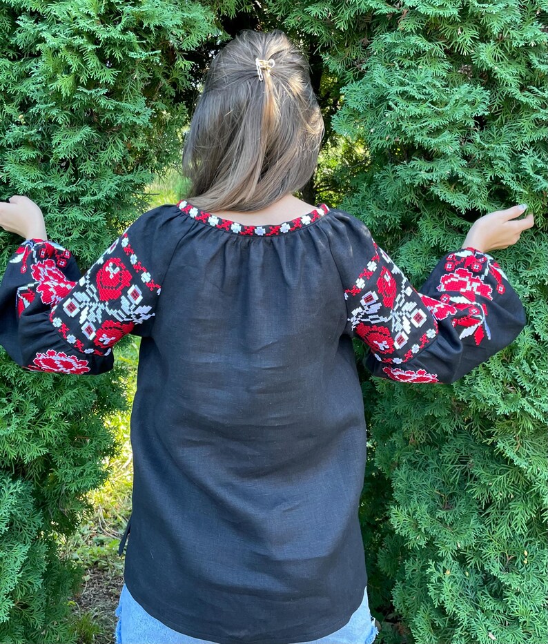Puede incluir: Una camisa de lino negra con flores bordadas rojas y blancas en las mangas. La camisa tiene un cuello redondo y un corte holgado.