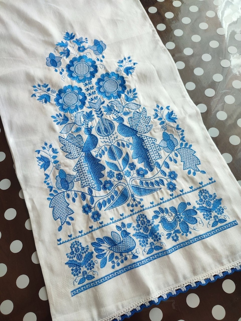 May include: White linen towel with intricate blue embroidery featuring floral and bird motifs. The design includes detailed flowers, leaves, and stylized birds, with a delicate blue lace trim along the edge. The towel is likely handmade.
