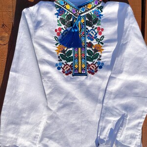 May include: White linen shirt with colorful embroidered floral pattern and blue tassel. The shirt has long sleeves with embroidered cuffs.