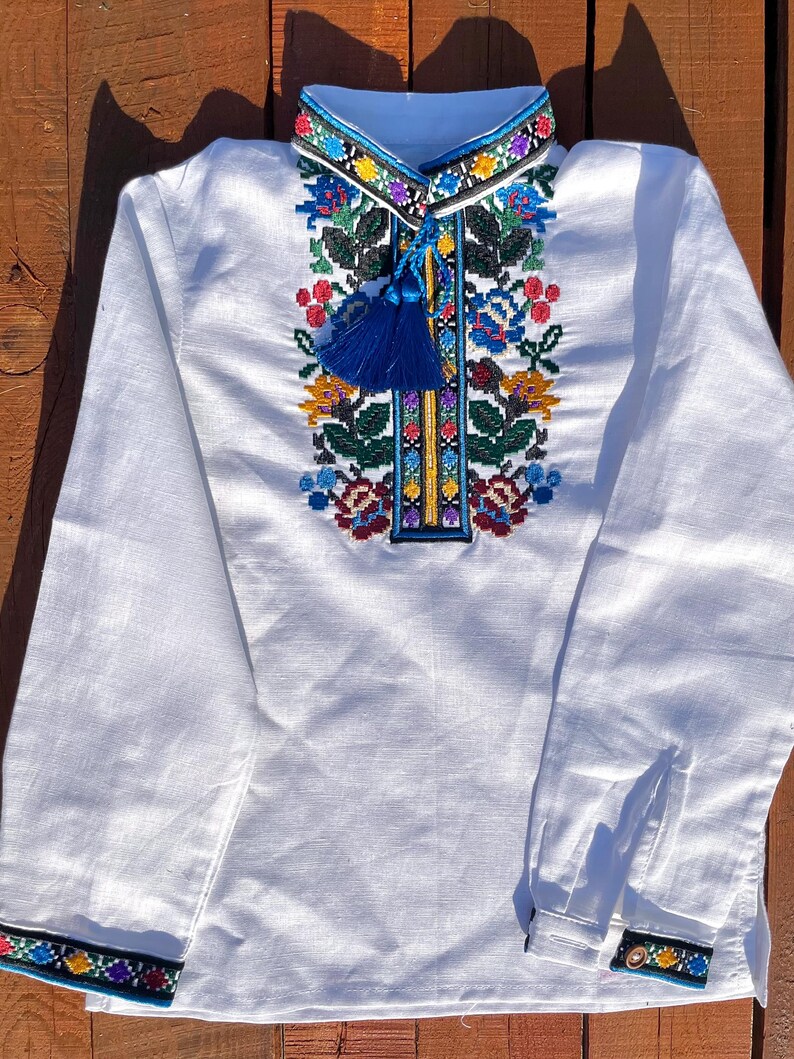 May include: White linen shirt with colorful embroidered floral pattern and blue tassel. The shirt has long sleeves with embroidered cuffs.