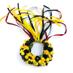 K&ouml;nnte beinhalten: Ein dekorativer Kranz mit gelben und schwarzen Blumen, Beeren und B&auml;ndern. Der Kranz ist mit kleinen gelben und schwarzen Blumen, gelben Beeren und schwarzen Beeren geschm&uuml;ckt. B&auml;nder in Gelb, Schwarz und Rot erstrecken sich von oben.