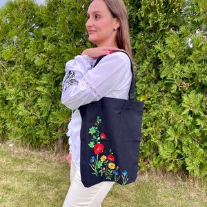 以下が含まれることがあります： 赤い、黄色い、青い、緑の花が刺繍された黒いトートバッグ。白いブラウスと白いパンツを着た人が持っています。