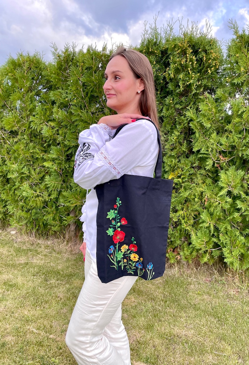 以下が含まれることがあります： 赤い、黄色い、青い、緑の花が刺繍された黒いトートバッグ。白いブラウスと白いパンツを着た人が持っています。