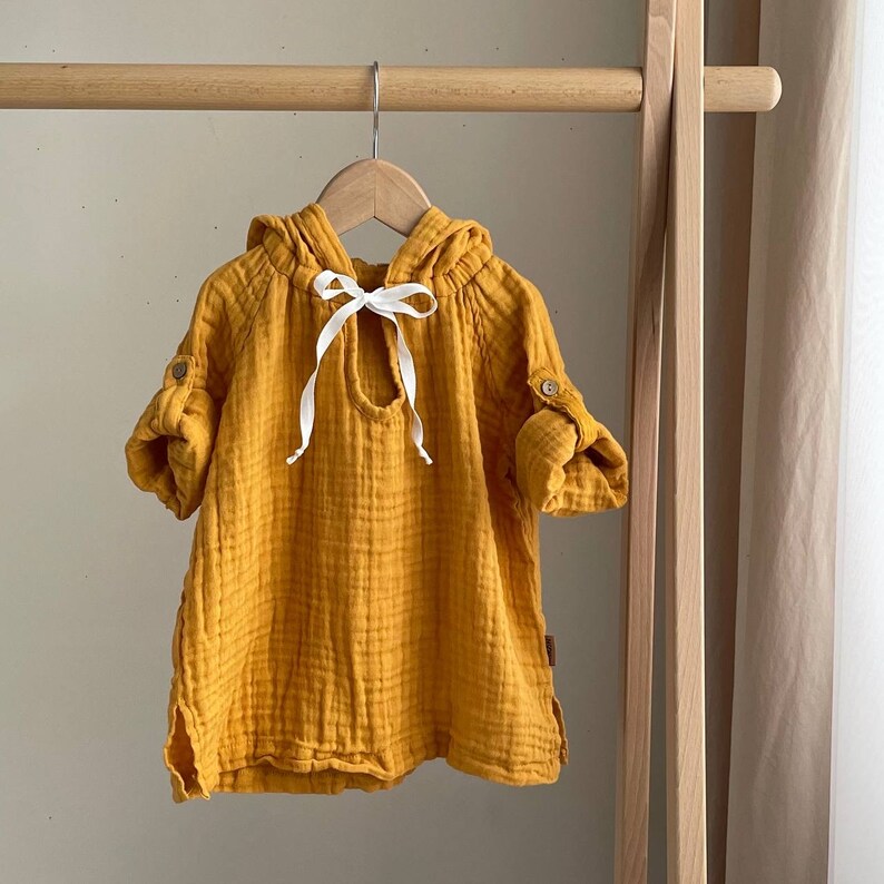 May include: A yellow hooded tunic with a white ribbon tie. The tunic is made of a soft, lightweight fabric and has a relaxed fit.