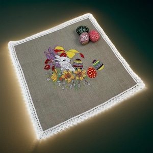 May include: Embroidered linen table runner with a white bunny rabbit, colorful flowers, and Easter eggs. The runner has a white lace trim.