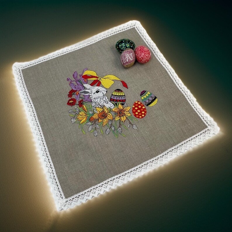 May include: Embroidered linen table runner with a white bunny rabbit, colorful flowers, and Easter eggs. The runner has a white lace trim.