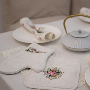 May include: Two embroidered linen pot holders with floral designs. One pot holder is shaped like a mitten and the other is a square. Both pot holders feature pink, red, and yellow flowers.