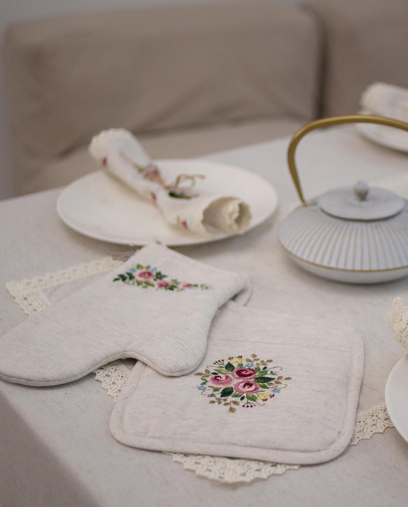 May include: Two embroidered linen pot holders with floral designs. One pot holder is shaped like a mitten and the other is a square. Both pot holders feature pink, red, and yellow flowers.