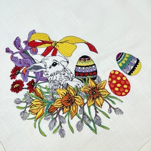 Op de afbeelding: Witte geborduurde keukendoek met een konijn, gele narcissen, paarse irissen en kleurrijke paaseieren. De tekst "Prettige Pasen" is in rood geborduurd.