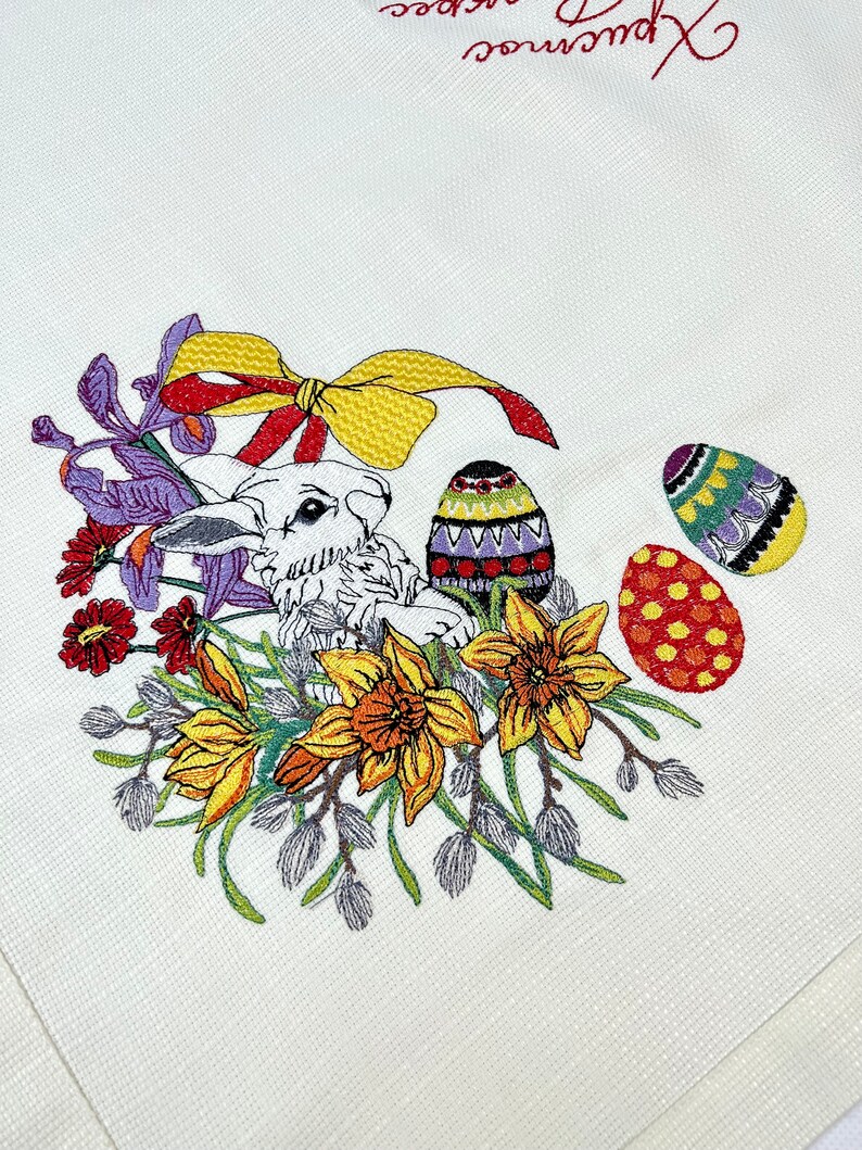 Op de afbeelding: Witte geborduurde keukendoek met een konijn, gele narcissen, paarse irissen en kleurrijke paaseieren. De tekst "Prettige Pasen" is in rood geborduurd.