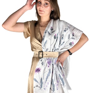 Op de afbeelding: Een vrouw draagt een beige jurk met een witte linnen sjaal met een bloemenpatroon. De sjaal heeft een paars en grijs bloemenpatroon met een bruine riem.