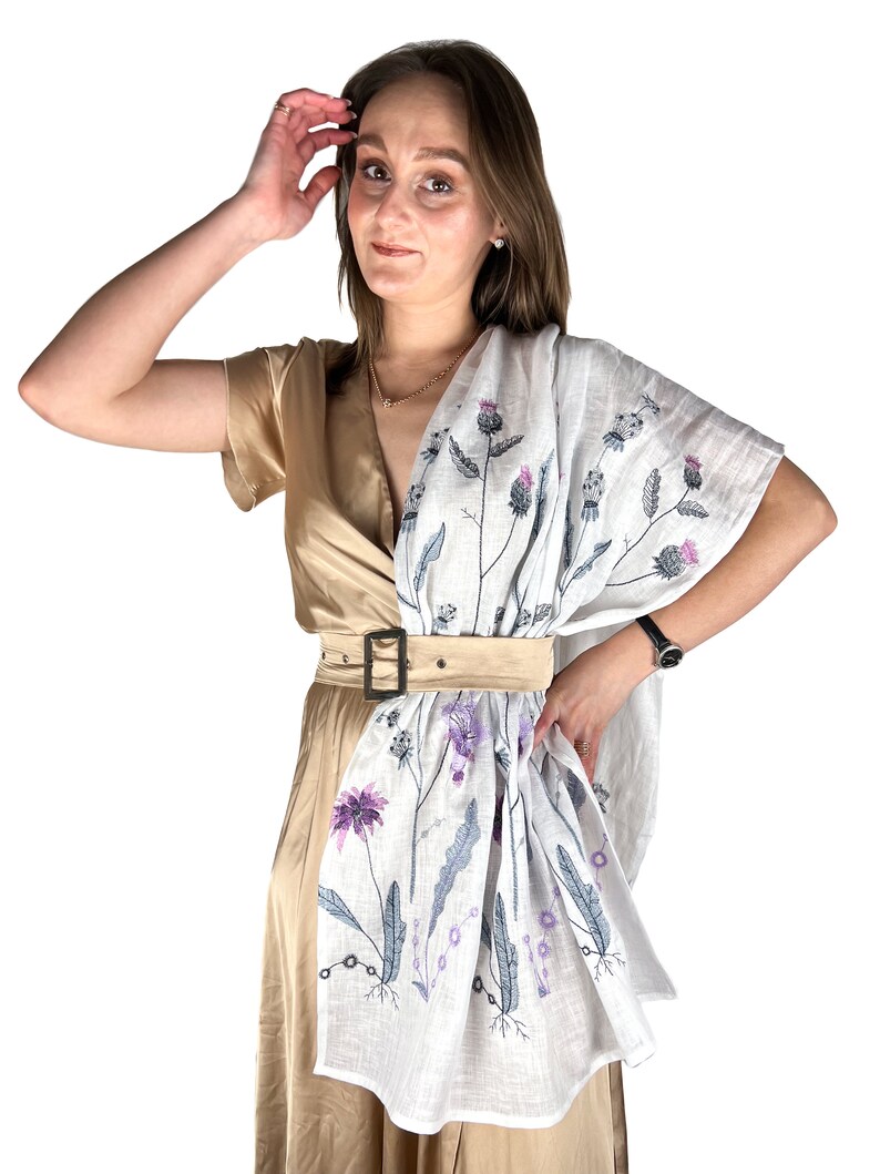 Op de afbeelding: Een vrouw draagt een beige jurk met een witte linnen sjaal met een bloemenpatroon. De sjaal heeft een paars en grijs bloemenpatroon met een bruine riem.