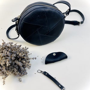 Puede incluir: Un bolso de hombro redondo de cuero negro con cierre de cremallera y una correa de cadena plateada. El bolso est&aacute; hecho de varias piezas de cuero cosidas juntas en un patr&oacute;n geom&eacute;trico. Tambi&eacute;n se incluye una peque&ntilde;a bolsa de cuero negro y un llavero de cuero negro con cierres plateados.