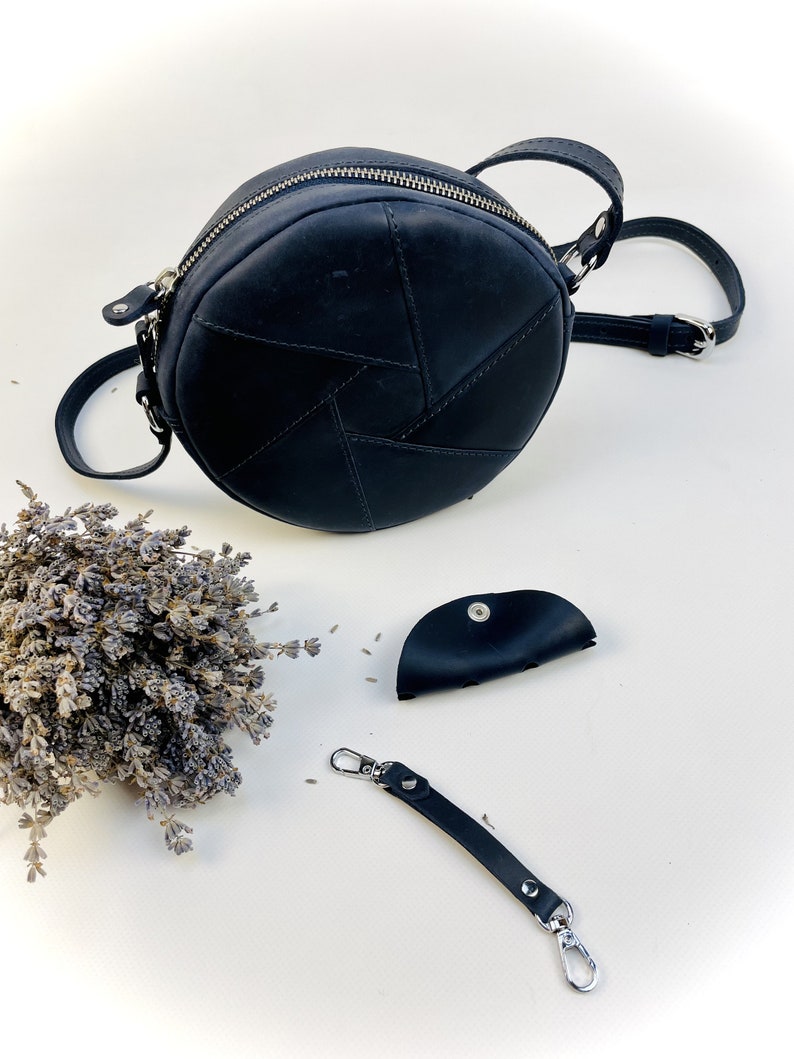 Puede incluir: Un bolso de hombro redondo de cuero negro con cierre de cremallera y una correa de cadena plateada. El bolso est&aacute; hecho de varias piezas de cuero cosidas juntas en un patr&oacute;n geom&eacute;trico. Tambi&eacute;n se incluye una peque&ntilde;a bolsa de cuero negro y un llavero de cuero negro con cierres plateados.