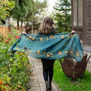 May include: A teal scarf with orange floral embroidery is held up in the air. The scarf is the focal point, with a woman walking away from the camera. The background includes a garden, a walkway, and a wooden building.