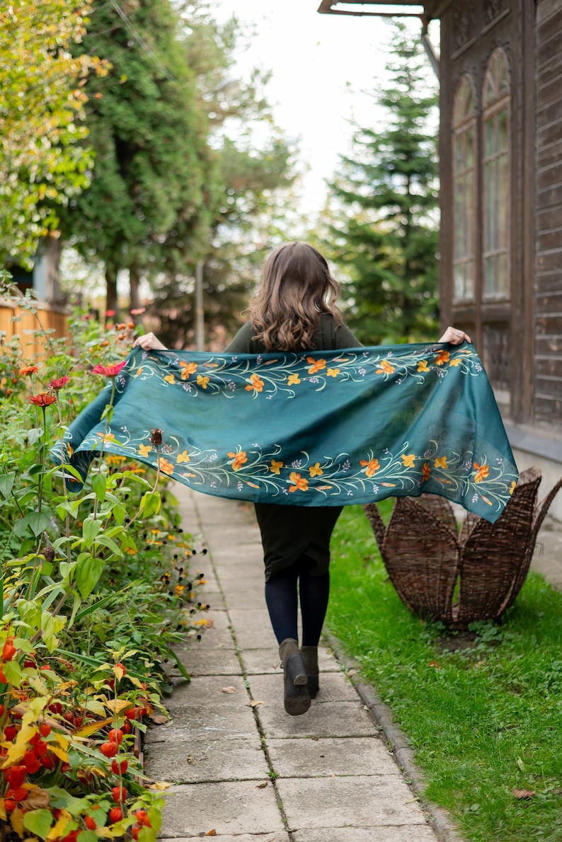 May include: A teal scarf with orange floral embroidery is held up in the air. The scarf is the focal point, with a woman walking away from the camera. The background includes a garden, a walkway, and a wooden building.