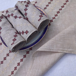May include: Beige linen table runner and napkins set. The runner and napkins feature embroidered geometric patterns in red and cream. Rolled napkins are placed on a blue and gold rimmed plate. The set is displayed on a white tablecloth.
