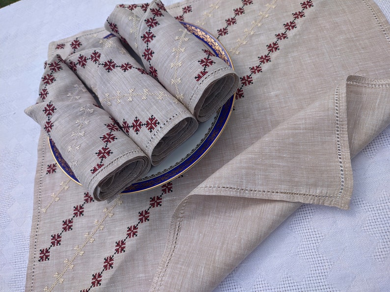 May include: Beige linen table runner and napkins set. The runner and napkins feature embroidered geometric patterns in red and cream. Rolled napkins are placed on a blue and gold rimmed plate. The set is displayed on a white tablecloth.