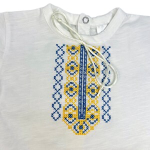 May include: White short-sleeved shirt with a blue and yellow embroidered design on the front. The embroidery features a geometric pattern.