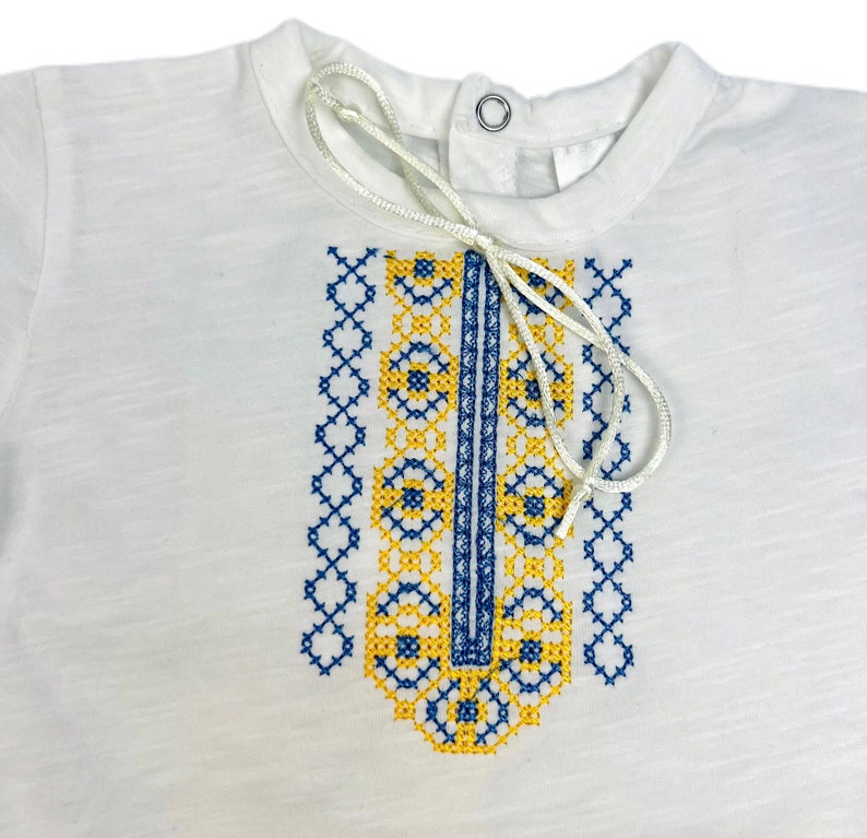 May include: White short-sleeved shirt with a blue and yellow embroidered design on the front. The embroidery features a geometric pattern.