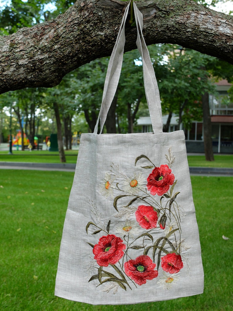 Op de afbeelding: Een linnen tas met geborduurde rode klaprozen en witte madeliefjes. De tas heeft een natuurlijke linnen kleur en hangt aan een tak.
