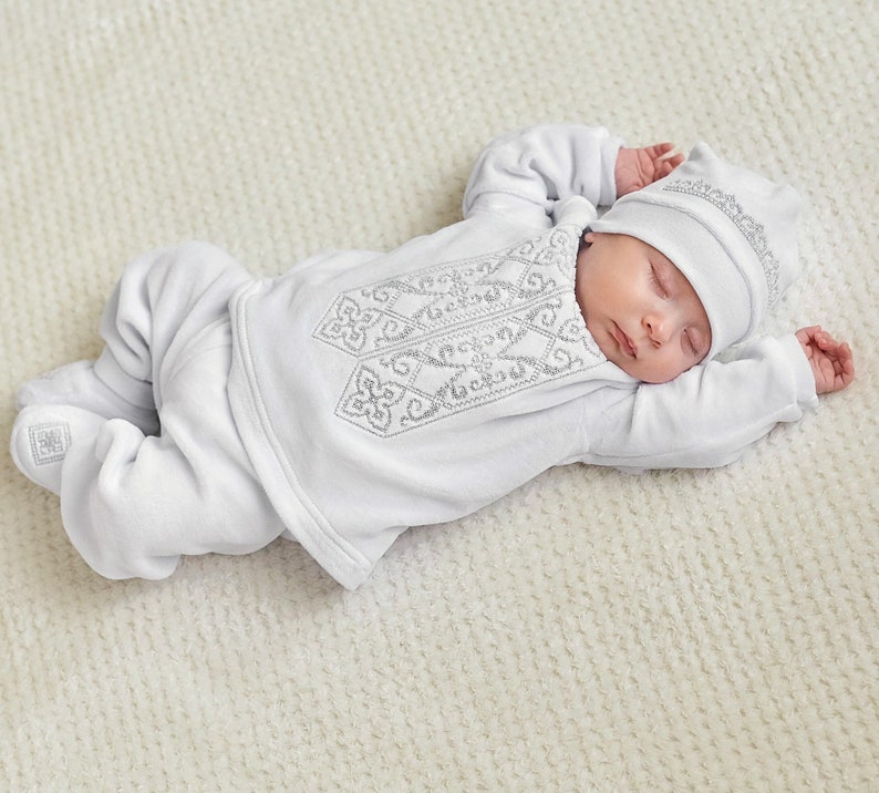 K&ouml;nnte beinhalten: Ein schlafendes Baby in einem wei&szlig;en Outfit mit silberner Stickerei. Das Outfit umfasst ein lang&auml;rmliges Oberteil, eine Hose und eine passende M&uuml;tze. Die Stickerei zeigt ein geometrisches Muster. Das Baby liegt auf einer wei&szlig;en, strukturierten Decke.