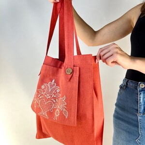Op de afbeelding: Een koraalkleurige tote bag met een groot vak aan de voorkant. Het vak heeft een wit geborduurd bloemenmotief. De tas heeft twee lange hengsels.