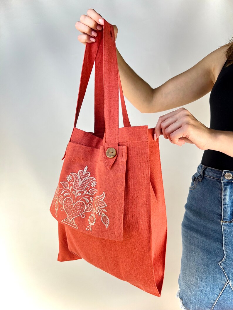 Op de afbeelding: Een koraalkleurige tote bag met een groot vak aan de voorkant. Het vak heeft een wit geborduurd bloemenmotief. De tas heeft twee lange hengsels.