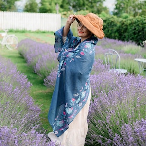 May include: A woman wearing a blue floral patterned shawl and a wide-brimmed brown hat stands in a field of lavender.