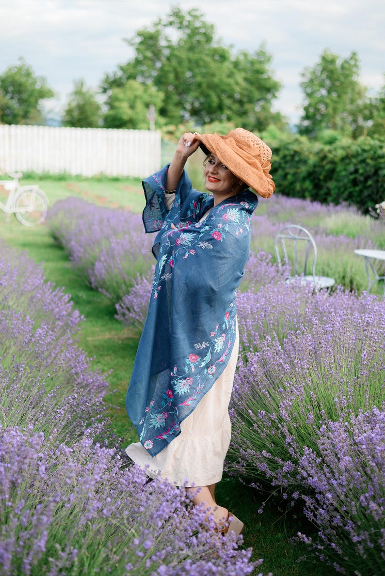 May include: A woman wearing a blue floral patterned shawl and a wide-brimmed brown hat stands in a field of lavender.