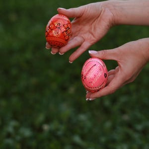 K&ouml;nnte beinhalten: Zwei handbemalte Ostereier, eines rot mit schwarzen und goldenen Mustern und eines rosa mit wei&szlig;en Mustern, werden in den H&auml;nden einer Person gehalten.