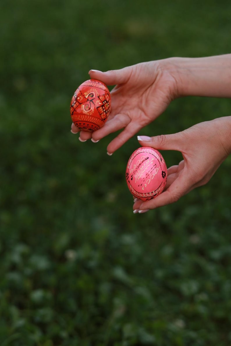 K&ouml;nnte beinhalten: Zwei handbemalte Ostereier, eines rot mit schwarzen und goldenen Mustern und eines rosa mit wei&szlig;en Mustern, werden in den H&auml;nden einer Person gehalten.