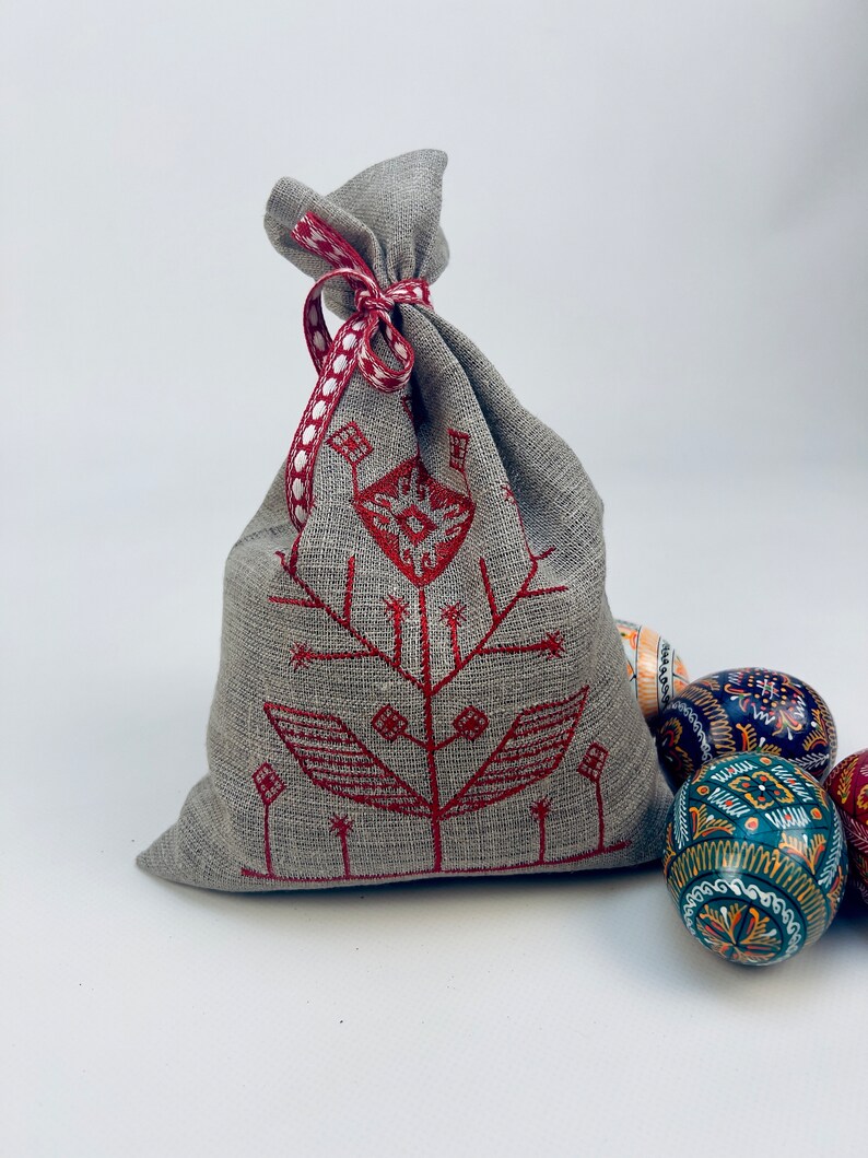 Pode incluir: Uma bolsa de linho cinza com um desenho floral vermelho bordado, amarrada com uma fita vermelha e branca. V&aacute;rios ovos de P&aacute;scoa decorados s&atilde;o vis&iacute;veis no fundo, mostrando um artesanato tradicional.