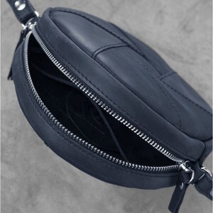 Puede incluir: Un bolso de hombro de cuero azul oscuro con forma circular y cierre de cremallera. El bolso tiene una correa y una cremallera plateada.