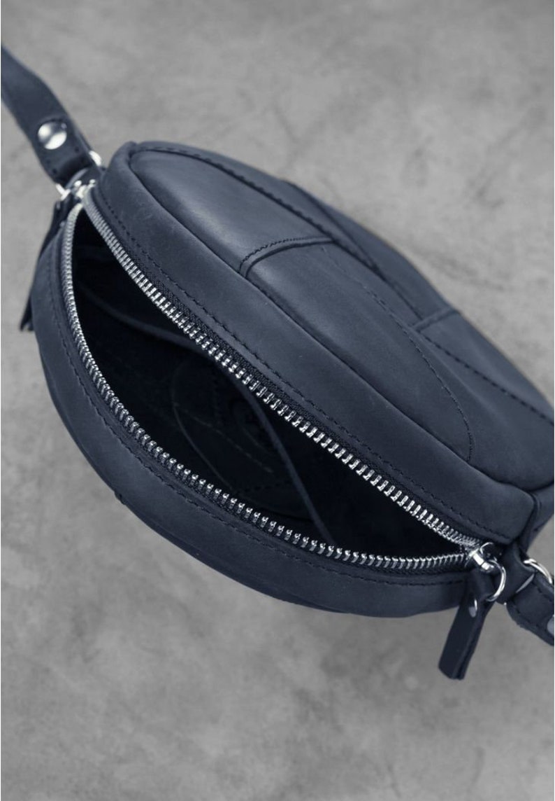 Puede incluir: Un bolso de hombro de cuero azul oscuro con forma circular y cierre de cremallera. El bolso tiene una correa y una cremallera plateada.
