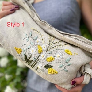 May include: A beige linen zippered pouch with a floral embroidery design. The embroidery features white flowers, yellow centers, and green leaves. The text "Style 1" is visible on the pouch.