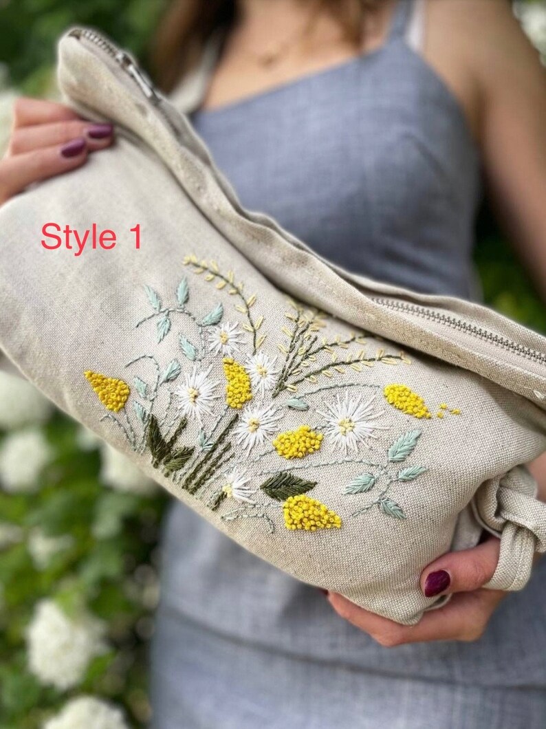 May include: A beige linen zippered pouch with a floral embroidery design. The embroidery features white flowers, yellow centers, and green leaves. The text "Style 1" is visible on the pouch.